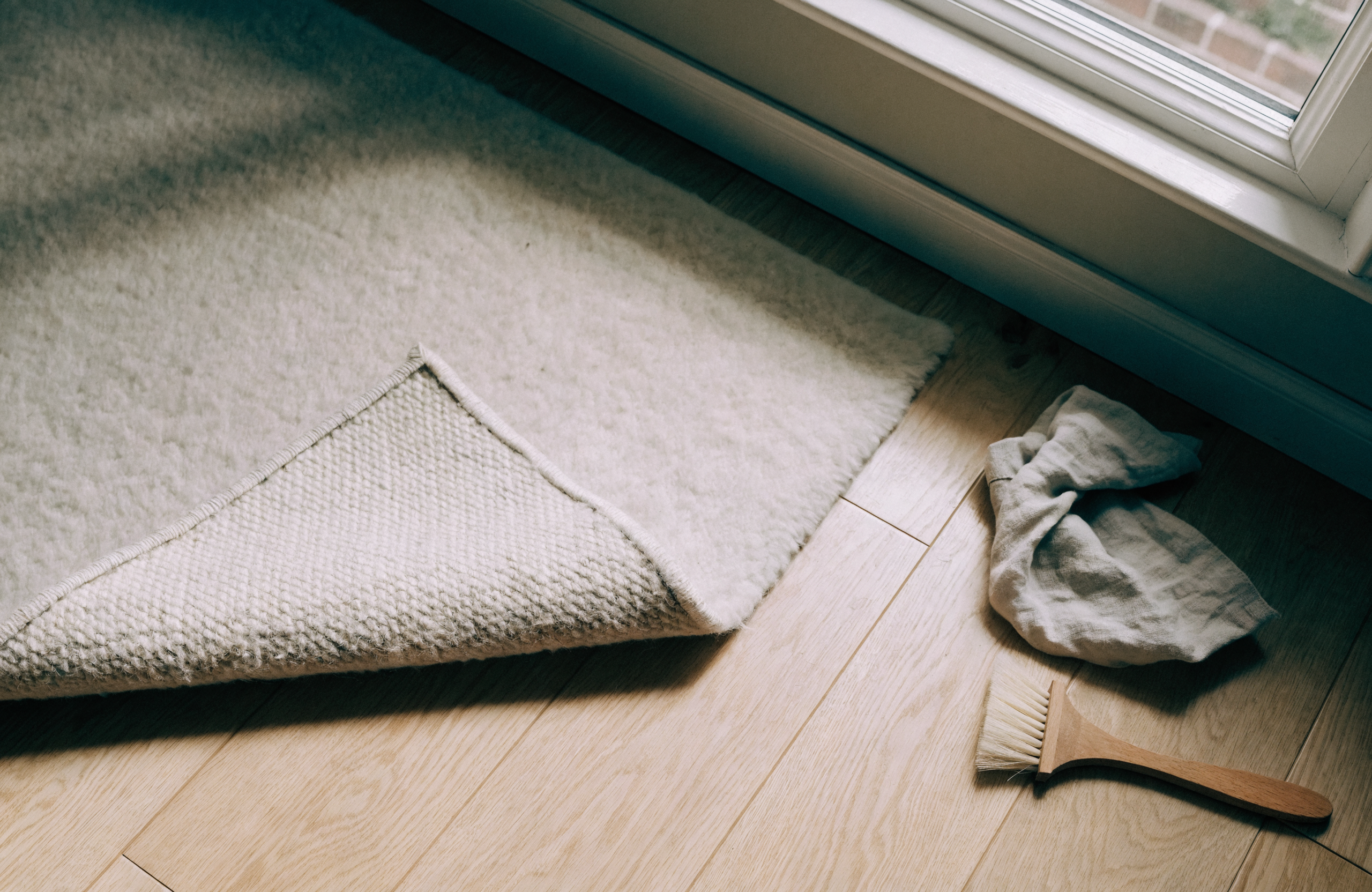 The corner of a cream wool rug lifted back on a wooden floor beside a soft brush and a folded cloth