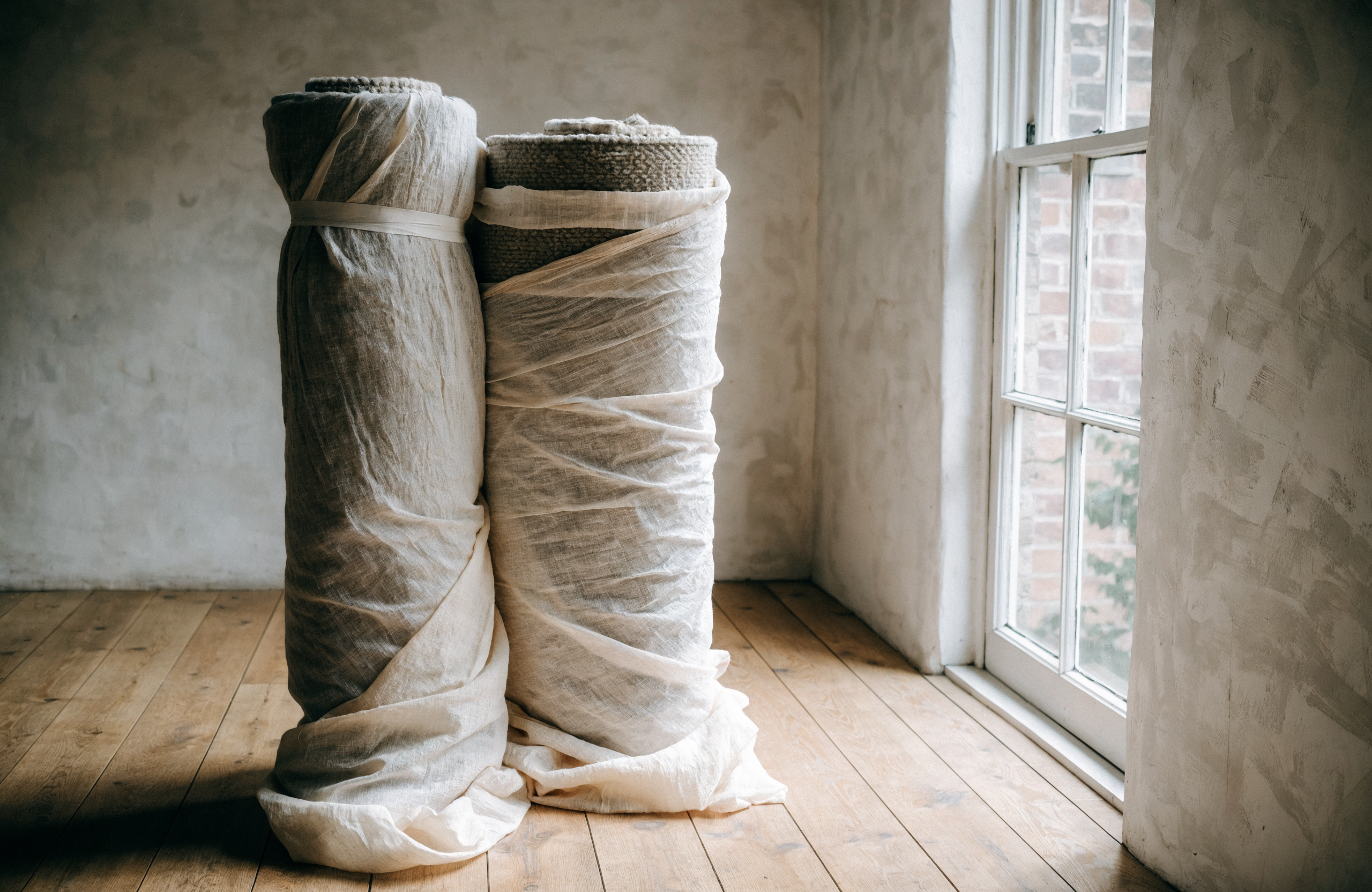 Two rugs rolled and wrapped in undyed cotton sheeting, stood in a bright empty room